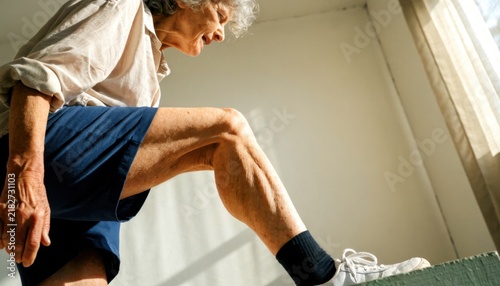 Senior functional fitness concept. Elderly man wearing shorts and sneakers exercising indoors with natural light streaming through a window.