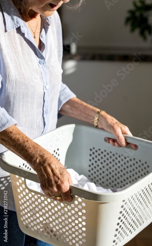 Senior functional fitness concept. Elderly woman holding a laundry basket filled with clothes, engaged in household chores in a bright room.