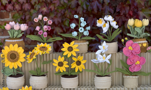 Many beautiful hand crafted crochet flowers in crocheted  pots on a tiered table. Leaves in background