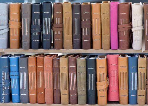 Rows of hand crafted leather bound books with various colors of covers on wood shelf.