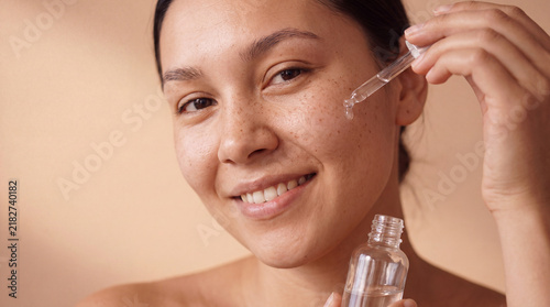 A young mixed-race woman holds an essence bottle with a soft smile for a beauty content post.