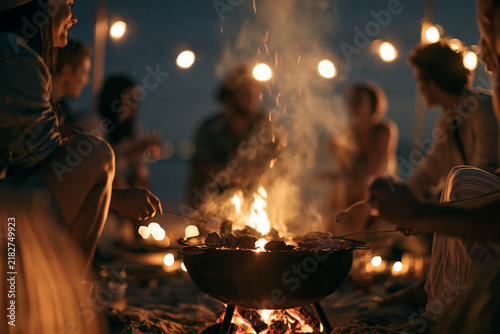 Group of friends enjoying outdoor barbecue at night, sitting around fire pit on sandy beach, warm lights in background, relaxed atmosphere, summer evening gathering, joyful and cozy mood