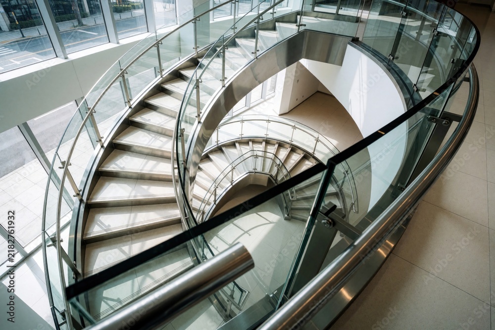 Fototapeta premium Modern spiral staircase with glass railings and steel frame ascends through a bright, airy building interior. This architectural marvel connects floors within a contemporary structure.