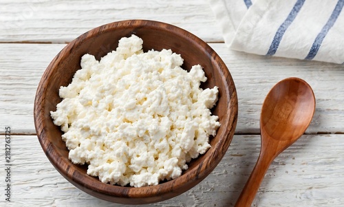 Fresh cottage cheese in a wooden bowl on a rustic white table with a wooden spoon.