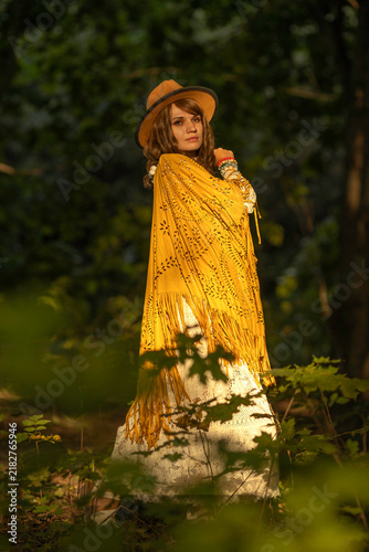 Cuadro en lienzo A young woman strolls calmly and casually through a field; she is wearing an open yellow shawl and a brown hat, with her arms spread wide
