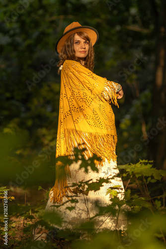 Fotografie A young woman strolls calmly and casually through a field; she is wearing an open yellow shawl and a brown hat, with her arms spread wide