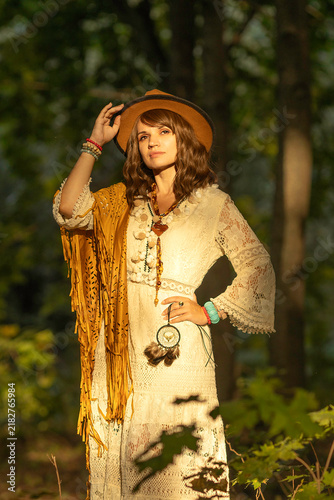 A young woman strolls calmly and casually through a field; she is wearing an open yellow shawl and a brown hat, with her arms spread wide. Boho style.
