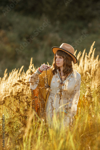 A young woman strolls calmly and casually through a field; she is wearing an open yellow shawl and a brown hat, with her arms spread wide. Boho style.