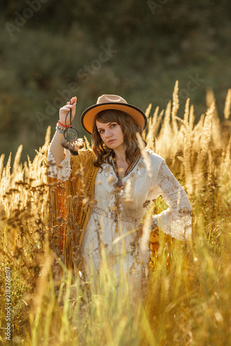 A young woman strolls calmly and casually through a field; she is wearing an open yellow shawl and a brown hat, with her arms spread wide. Boho style.