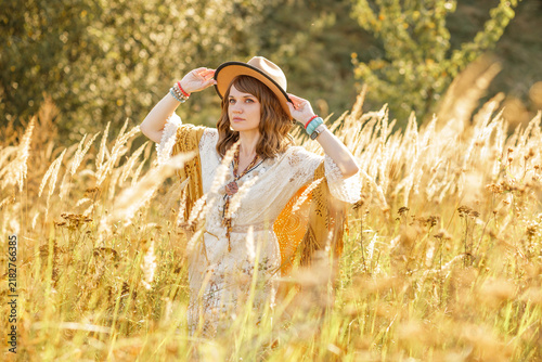 A young woman strolls calmly and casually through a field; she is wearing an open yellow shawl and a brown hat, with her arms spread wide. Boho style.