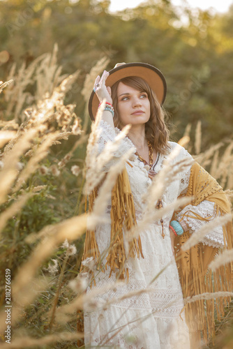 A young, fair-skinned woman strolls calmly and casually through a field; she is wearing an open yellow shawl and a brown hat, with her arms spread wide. Boho style.