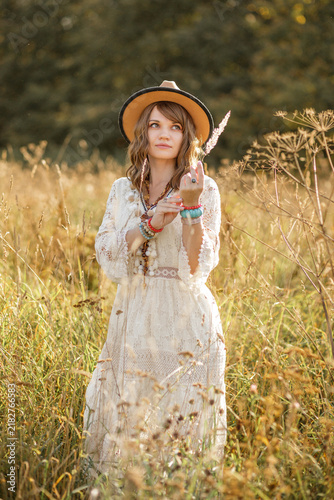 A young, fair-skinned woman strolls calmly and casually through a field; she is wearing an open yellow shawl and a brown hat, with her arms spread wide. Boho style.