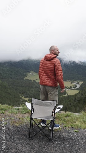 Man sits in a portable camping chair above a mountain valley, relaxing and enjoying the scenic cloudy landscape.