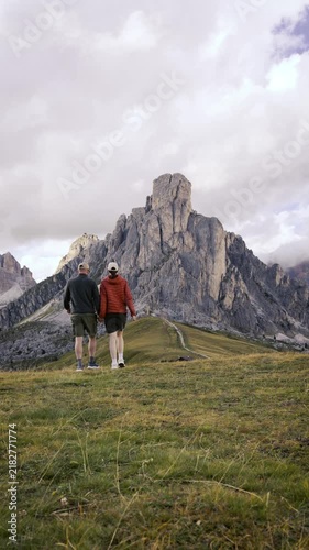 A young couple hiking together in the Dolomites, Italy, walking through an alpine meadow and relaxing on the grass while enjoying a dramatic mountain landscape and scenic nature view.
