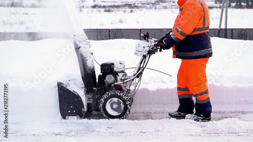 Worker clears snow with heavy-duty snow blower. Bright orange suit signals safety and visibility. Snow sprays forcefully into air behind machine. Scene shows winter cleanup effort in progress