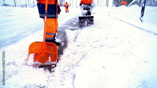 Worker clears snow with bright orange shovel. Snow plows follow behind, clearing paths. Team works in cold, snowy conditions. Essential winter maintenance scene. Urban or rural road clearing footage
