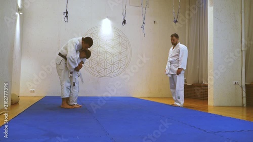 Two men in white uniforms bow respectfully. One kneels, eyes closed, showing deep focus. Another stands, observing with calm patience. Bright light shines behind them, illuminating the blue mat