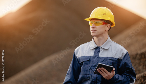 Industrial worker with tablet and yellow safety helmet in sand quarry at sunset for site inspection