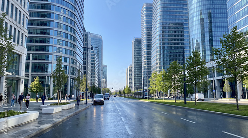 Modern glass corporate skyscrapers flank wide urban street with morning light and greenery.
