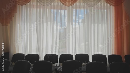 Back view of empty chairs in an empty conference hall. The wind gently moves the thin white curtains, showing the sky outside the window