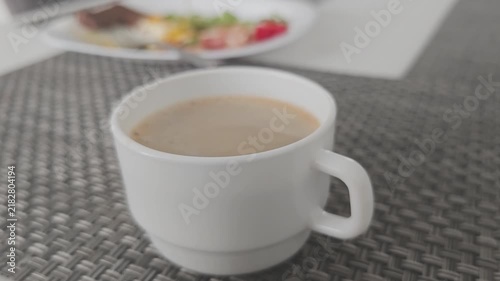 Close up of a warm cup of coffee with milk on a placemat. Morning beverage steaming on a table with a plate of food in the background