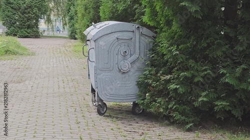 Large gray metal dumpster on wheels standing on a paved pathway next to a green hedge. Outdoor garbage collection and waste management concept