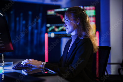Businessman and businesswoman standing at a glowing AI brain wall behind desk monitors raise a stylus to highlight a detail during a product launch rehearsal in a dark high tech auditorium