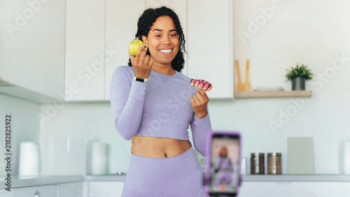 Smiling African American Woman Holding An Apple And A Doughnut In A Modern Kitchen  Recording A Video. Cheerful Female Influencer Promoting Healthy Eating And Lifestyle With Visual Engagement.