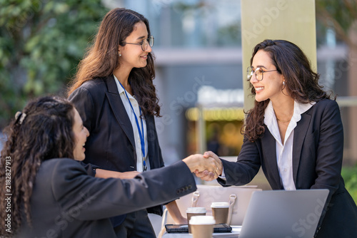 A diverse team of three Hispanic businesswomen in dark suits  smiling with mutual respect  welcome a new partner to the table reaching across the table to shake hands.