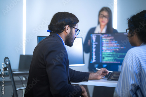 A male software engineer in a suit checking project data on a tablet device  at a long desk with code monitors against a bright window  side profile view of software development.
