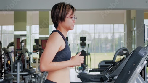 Active Asian woman in her forties wearing glasses and a black sports bra runs on a treadmill inside a modern brightly lit gym. She is focused on her cardio workout to maintain a healthy lifestyle.