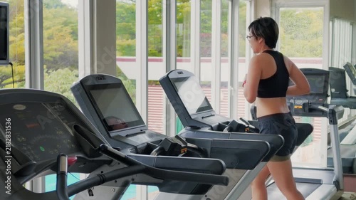 Wide shot of an active Asian woman in her forties running on a treadmill in a bright modern gym. She wears a black sports bra, enjoying daily cardio exercise by large windows with natural daylight.