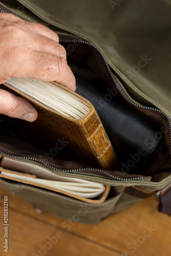 Man taking a vintage leather journal out of a green canvas messenger bag satchel. On a wooden floor background.