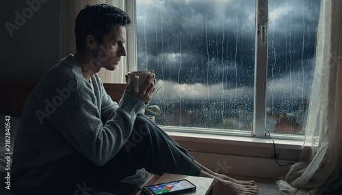 Man Sitting by Window Watching Storm and Rain Outside, Holding Coffee Mug While Checking Weather App on Smartphone, Weather Anxiety Concept