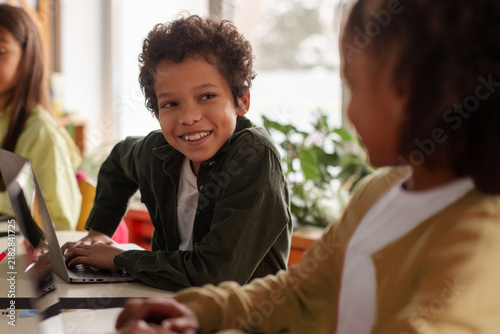фотографія Smiling mixed-race elementary school boy looking up from his laptop at classmate