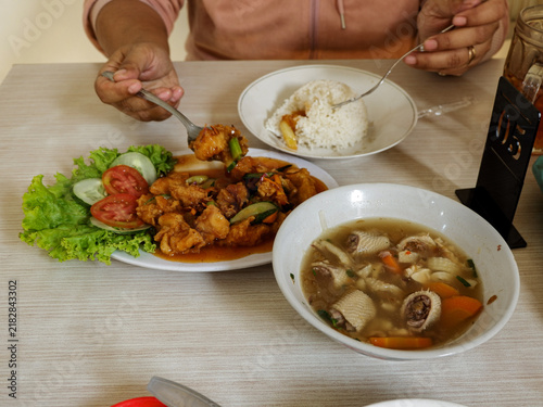 Canvastavla Person holding cutlery while enjoying a dining table setup featuring chicken with sauce, vegetable soup, and white rice