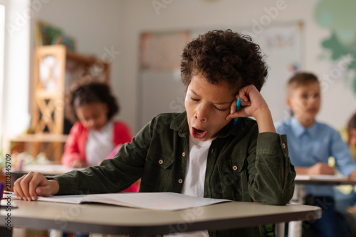 Bild auf Leinwand Schoolboy yawning while studying in classroom, expressing boredom during lesson