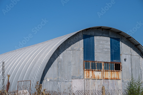 a large, weathered corrugated metal Quonset hut standing behind a wire fence in an overgrown field of dry grass under a clear blue sky.