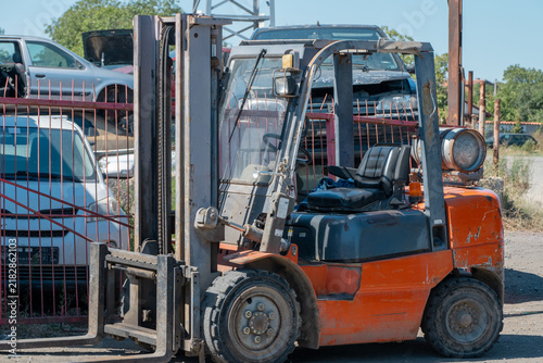 Photography a weathered orange forklift stands parked near a red metal fence in a salvage yard filled with dismantled cars