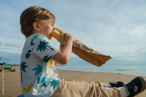 Cuadro en lienzo Happy young boy sits on sandy beach, savoring snack from brown paper bag with great enthusiasm