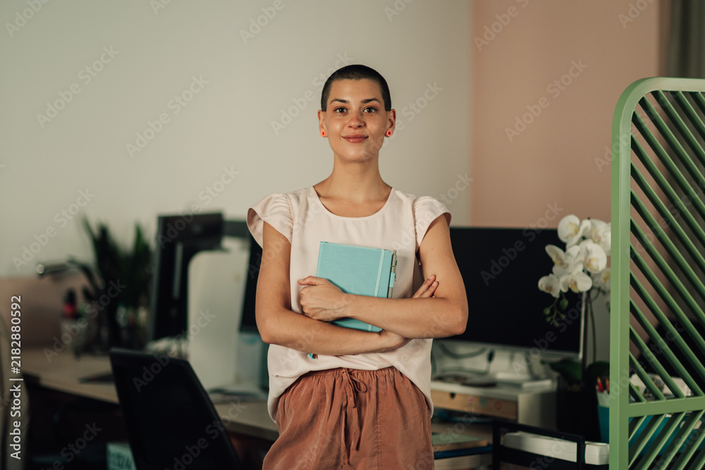 Obraz premium Confident young woman with short hair holding a diary, smiling and standing in a modern office workplace