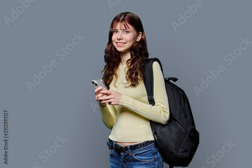 Smiling young female student with smartphone and backpack looking at camera on grey background