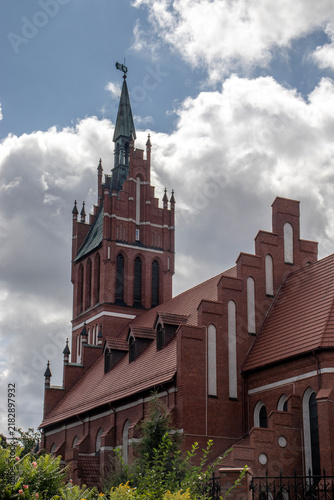A historic brick church building with a tall, narrow tower and a pointed spire, photographed from below at an angle against a blue sky with large white clouds. The reddish-brown brick walls