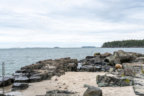 Roque Bluffs State Park rugged dark rocky coastline and sandy beach shoreline overlooking calm Atlantic ocean waters under an overcast sky in Maine.