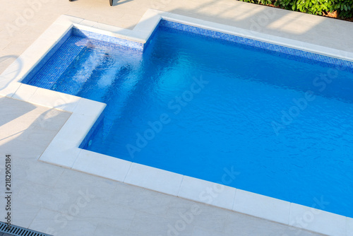 Фотография A clear swimming pool surrounded by white stone deck and green plants in a sunny