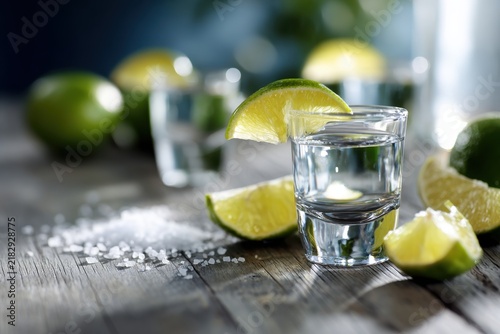Clear drink shot glass garnished with fresh lime wedge on rustic wooden surface surrounded by coarse salt and lime slices in soft natural light