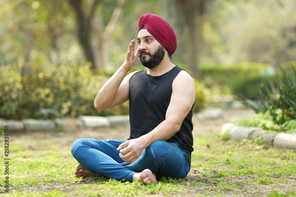 Fototapeta premium Concept of young sikh man yoga at park.. Cheerful sardar in lotus pose sitting on green grass. Concept of calm and meditation.. young man practicing yoga in park. 