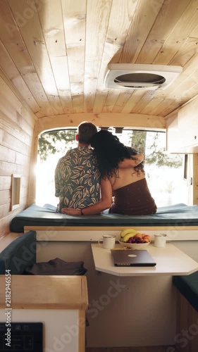 Young couple sitting on a bed inside a recreational vehicle. Man and woman looking out the back window at the forest enjoying their vacation. Vertical