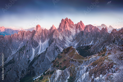 First light turns a wall of jagged limestone spires deep pink above steep scree slopes and a winding mountain trail