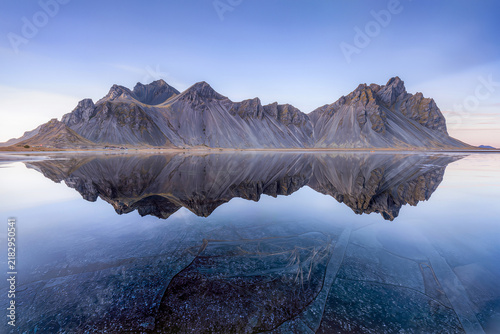 A serrated coastal mountain mirrors perfectly in a still lagoon fringed with cracked sheets of clear ice at blue hour
